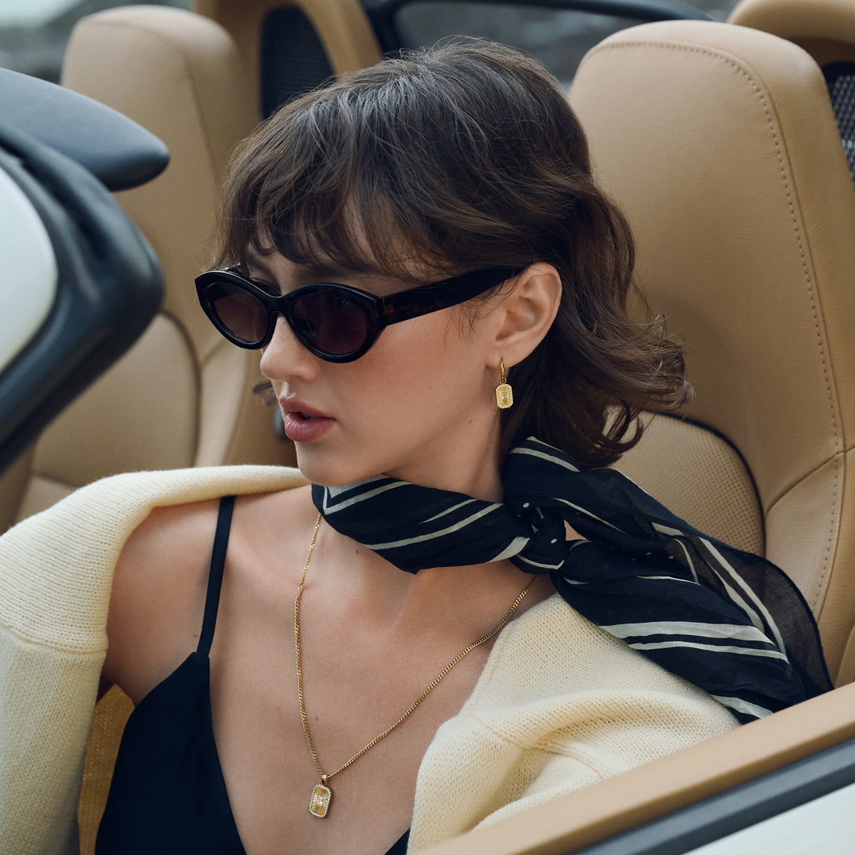 Close-up lifestyle shot of a woman seated in a car, wearing gold hoop earrings with a rectangular sunburst pendant and a matching gold chain necklace, styled with sunglasses, a striped scarf and a cream knit.
Photo lifestyle en gros plan d'une femme assise dans une voiture, portant des boucles d'oreilles anneaux dorées à pendentif rectangulaire rayonnant et un collier à chaîne dorée assorti, agencés avec des lunettes de soleil, un foulard rayé et un tricot crème.