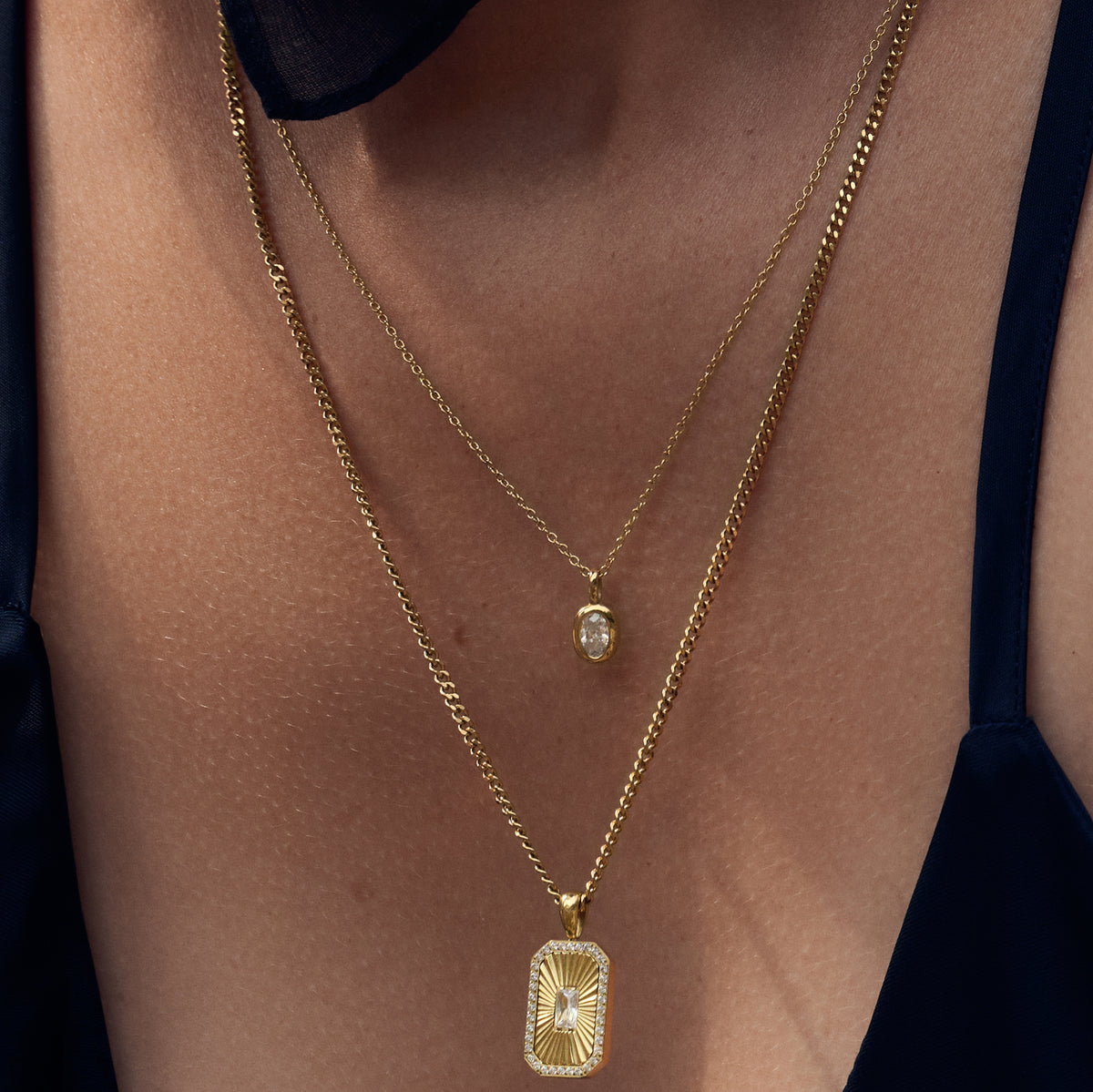 Close-up of a model wearing two layered gold necklaces, one with a small oval stone pendant and the other with a rectangular sunburst pendant. Waterproof, stainless steel and allergy free. By MIAJWL.
Gros plan d'un mannequin portant deux colliers dorés superposés, l'un à petit pendentif ovale serti et l'autre à pendentif rectangulaire rayonnant. Résistant à l'eau, en acier inoxydable et hypoallergénique. Par Mia Bijoux.
