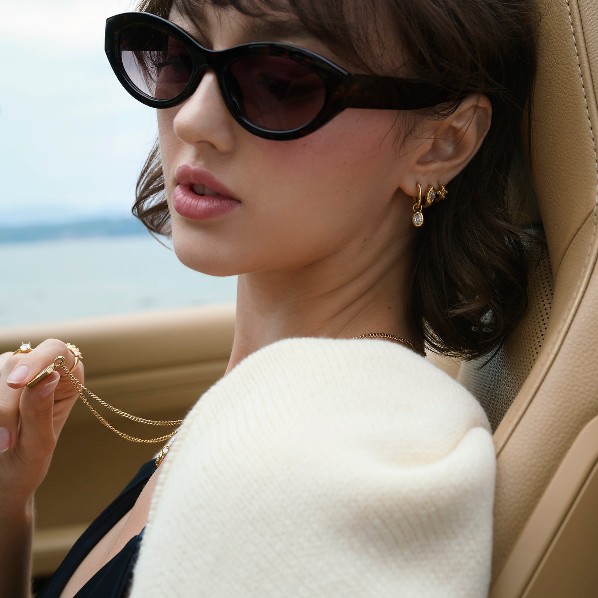 Close-up lifestyle shot of a woman seated in a car by the water, wearing layered gold earrings set with clear stones, a fine gold chain necklace and gold rings, styled with sunglasses and a cream knit.

Photo lifestyle en gros plan d'une femme assise dans une voiture au bord de l'eau, portant des boucles d'oreilles dorées superposées serties de pierres transparentes, un collier à chaîne fine dorée et des bagues dorées, agencés avec des lunettes de soleil et un tricot crème.