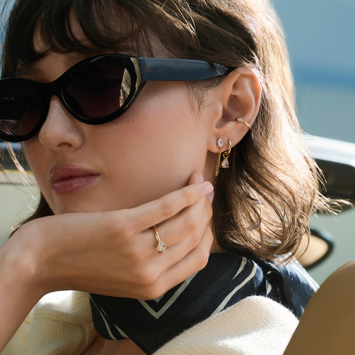 A close-up lifestyle shot of a woman wearing a gold pear-cut stone ring with matching gold earrings and ear cuff, styled with sunglasses, a navy scarf and a cream knit.

Photo lifestyle en gros plan d'une femme portant une bague dorée sertie d'une pierre taille poire, agencée avec des boucles d'oreilles dorées assorties et une manchette d'oreille, des lunettes de soleil, un foulard bleu marine et un tricot crème.