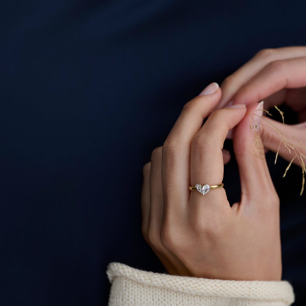 Close-up of a model's hands wearing a slim gold ring set with two pear-cut clear stones, styled with a cream knit sleeve against a dark navy background. Nickel-free, waterproof & tarnish-resistant.

Gros plan sur les mains d'un mannequin portant une bague fine dorée sertie de deux pierres transparentes taille poire, agencée avec une manche en tricot crème sur fond bleu marine foncé. Bague en acier inoxydable sans nickel, résistante à l'eau et anti-ternissement.