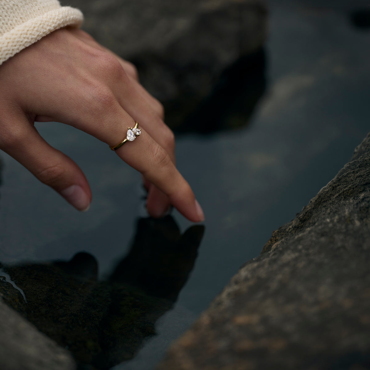 A close-up lifestyle shot of a hand wearing a delicate gold ring with a small clear gemstone, resting near dark water and rocks.

Photo lifestyle en gros plan d'une main portant une bague fine dorée sertie d'une petite pierre transparente, posée près d'une eau sombre et de rochers.